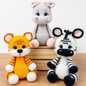 Op de afbeelding: Drie handgemaakte gehaakte knuffels, waaronder een gele en witte tijger met zwarte strepen, een lichtgrijs nijlpaard met roze oordetails en een wit-zwart gestreepte zebra, allemaal voorzien van grote zwarte veiligheidsogen en stiksels.