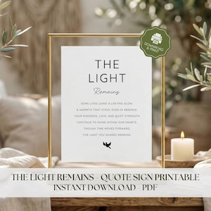 May include: A framed print with the words "THE LIGHT REMAINS" in black font. The print includes a poem about lasting memories and a small bird graphic. A candle and greenery are in the background.
