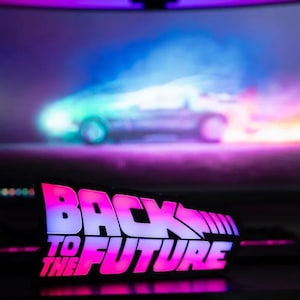 May include: Illuminated "Back to the Future" sign with a pink and white color scheme. The sign is in front of a computer monitor displaying a blurred image of the DeLorean time machine. The overall theme is retro sci-fi.