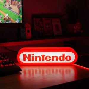 Könnte beinhalten: Ein rot-weißes Nintendo-Logo-Licht auf einem Schreibtisch, mit Tastatur und Maus. Ein Computermonitor zeigt eine Videospielszenen. Das Thema ist Gaming und Unterhaltung.