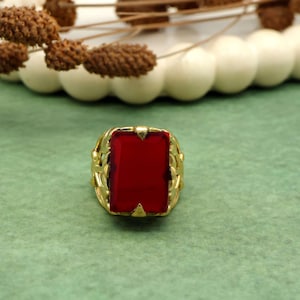 Puede incluir: Un anillo dorado con una gran gema roja rectangular. El anillo tiene un diseño ornamentado, similar a un tejido, alrededor de la gema. El anillo está sobre un fondo verde con elementos decorativos.