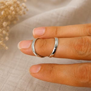 Könnte beinhalten: Ein Paar polierte Silberringe mit einem glatten, abgerundeten Banddesign. Die Ringe zeichnen sich durch eine reflektierende metallische Oberfläche und eine schlichte, klare Ästhetik aus. Jeder Ring ist aus einem massiven, durchgehenden Stück silberfarbenem Metall gefertigt, ohne sichtbare Gravuren oder Steine.