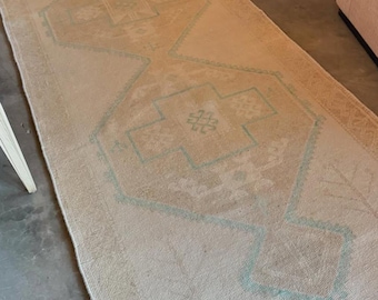 May include: A long rectangular runner rug featuring a light beige base color with faded geometric patterns in soft teal and muted tan tones. The design includes three large diamond-shaped medallions with intricate linear motifs and fringed edges.