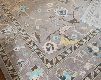 May include: A large area rug featuring a taupe background with a repeating floral and vine pattern in shades of light blue, yellow, brown, and cream. The rug has a wide border with geometric motifs and white fringe along the short edge.