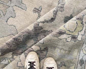 May include: A pair of white leather trainers with black laces featuring text, worn on a textured area rug. The rug has a light beige base with a faded, intricate floral pattern in shades of grey, charcoal, and muted yellow, showing a folded corner.