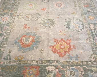 May include: A rectangular area rug featuring a light gray background with a wide, dark green border. The rug is decorated with scattered floral and geometric motifs in shades of orange, blue, yellow, and soft pink throughout the center field.