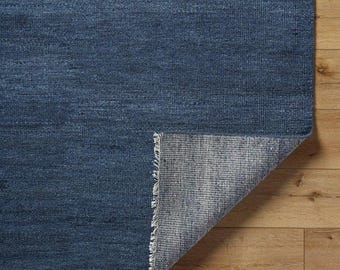 Puede incluir: Una alfombra azul con una superficie texturizada. La esquina de la alfombra está doblada hacia atrás, revelando la parte inferior con un patrón tejido gris y blanco. La alfombra está colocada sobre un suelo de madera.