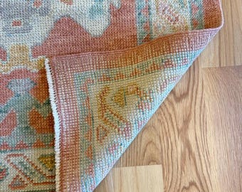 May include: A woven area rug featuring a geometric pattern in shades of peach, sage green, cream, and muted yellow. The rug has a folded corner revealing the underside, showcasing a textured pile and a white fringe edge along the side border.
