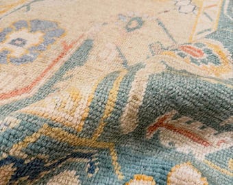 May include: A woven area rug featuring a cream-colored base with a detailed floral and geometric pattern in shades of teal, soft blue, muted yellow, and coral. The rug has a textured, knotted pile surface with visible fibers and a folded edge.