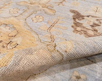 May include: A hand-knotted area rug featuring a light blue background with intricate floral and botanical patterns in shades of cream, beige, and brown. The rug has a dense, textured pile with a visible fold that reveals the thick construction.