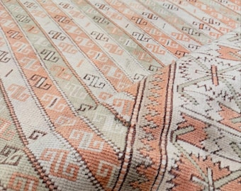 May include: A woven rug featuring a repeating pattern of peach and sage green geometric shapes on a cream background. The design includes alternating stripes of angular motifs and a border with a serrated edge, showcasing a textured pile surface.