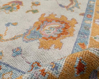 May include: A rectangular area rug featuring a cream-colored field with a repeating floral pattern in shades of orange, light blue, and lavender. The rug has a wide, textured orange border with blue and white accents and a visible pile texture.