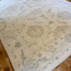 May include: A large area rug featuring a cream-colored base with intricate, faded floral and vine patterns in soft shades of light blue and beige. The design includes symmetrical medallion motifs and a decorative border, creating a textured appearance.