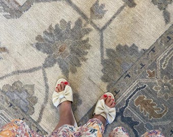 Peut inclure: Une paire de sandales à enfiler couleur crème dotées d'un nœud proéminent sur la bride supérieure, portées avec une robe à motifs floraux dans des tons orange, violet et bleu, mettant en valeur un design textile vibrant et coloré.