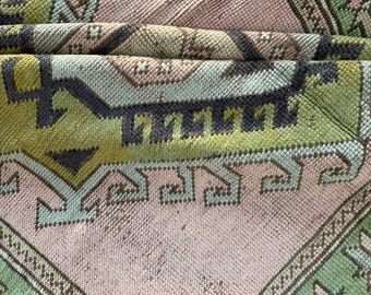 Peut inclure: Un tapis tissé présentant un motif géométrique dans des tons de vert sauge, rose pâle et gris anthracite foncé. Le textile affiche un motif répétitif avec des bordures angulaires et des formes en losange, avec une texture dense et épaisse.
