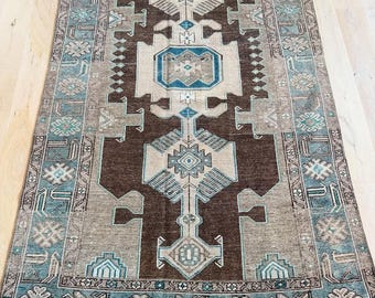 May include: A rectangular area rug featuring a dark brown central field with geometric medallions in beige and teal. The border displays a repeating pattern of floral motifs and angular lines in shades of teal, brown, and cream on a neutral background.