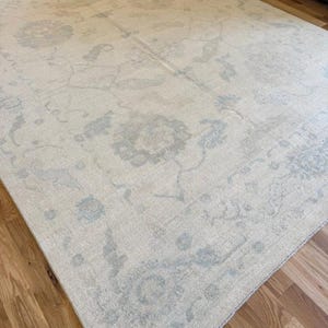 May include: A large rectangular area rug featuring a cream-colored base with a faded, intricate floral and vine pattern in soft shades of light blue and grey. The rug has a low-pile texture and covers a portion of a light brown wooden floor.