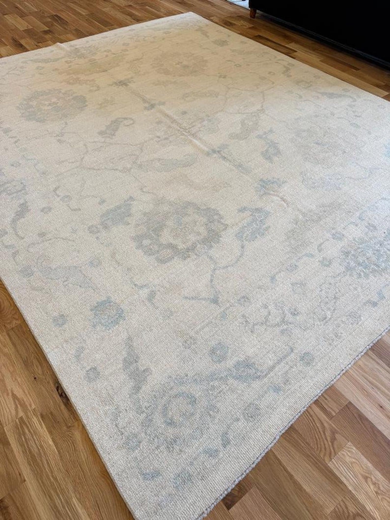 May include: A large rectangular area rug featuring a cream-colored base with a faded, intricate floral and vine pattern in soft shades of light blue and grey. The rug has a low-pile texture and covers a portion of a light brown wooden floor.