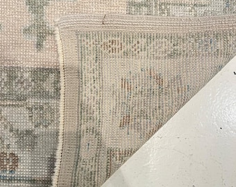 May include: A woven area rug featuring a folded corner that reveals the underside construction. The rug displays a pattern of muted sage green, soft pink, and beige floral motifs with a detailed border, showcasing the dense pile and fabric texture.