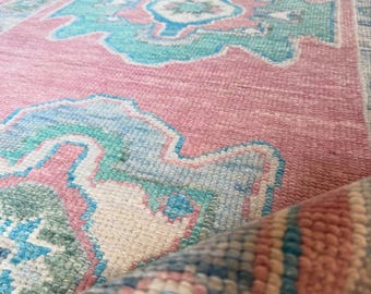 May include: A long runner rug featuring a pink base with repeating geometric medallion patterns in shades of turquoise, light blue, and cream. The rug has a textured, woven pile and a rolled edge that reveals a striped pattern of pink and blue.