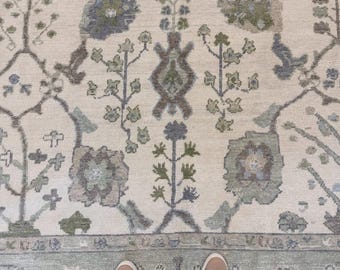 Peut inclure: Un grand tapis avec un fond couleur crème orné d'un motif floral et végétal complexe dans des tons sourds de vert sauge, de bleu poussiéreux et de gris anthracite. Le design présente des motifs botaniques symétriques et une large bordure.