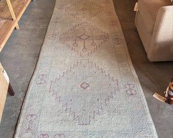 May include: A long, rectangular runner rug featuring a light beige base color with a repeating pattern of large, geometric diamond shapes in a muted purple hue. The rug has a textured, woven surface and a fringed edge along the short ends.