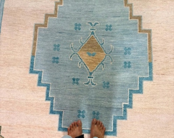 Peut inclure: Un grand tapis avec un médaillon géométrique central bleu clair et un losange beige au milieu. Le tapis présente un fond crème avec des bordures en escalier bleu clair et de petits motifs en forme de croix bleus dispersés sur la surface.