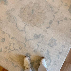 May include: A large area rug featuring a cream-colored base with a faded, light blue floral and vine pattern. The rug has a textured, woven appearance and covers a significant portion of the floor, extending beneath a pair of white sneakers.