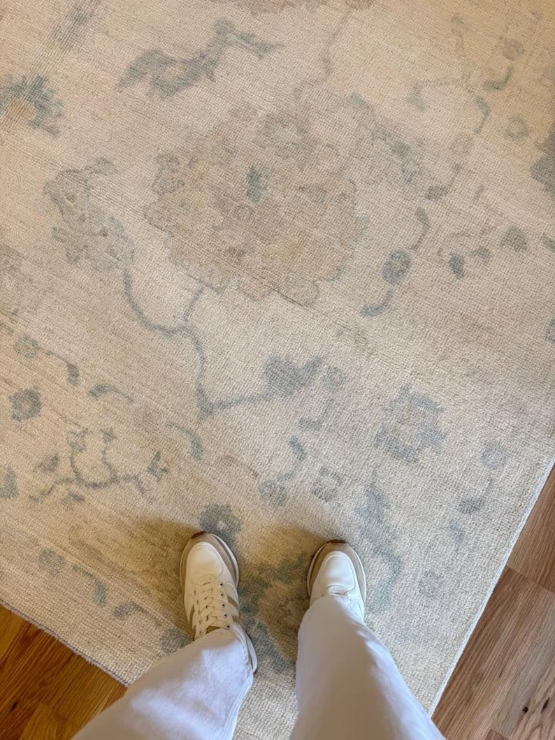 May include: A large area rug featuring a cream-colored base with a faded, light blue floral and vine pattern. The rug has a textured, woven appearance and covers a significant portion of the floor, extending beneath a pair of white sneakers.