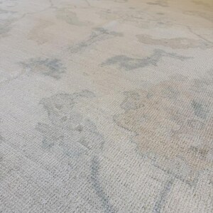 May include: A large area rug featuring a cream-colored base with a subtle, faded pattern in soft shades of light blue and beige. The rug has a low-pile, textured surface with an intricate, repeating design that appears worn or distressed.