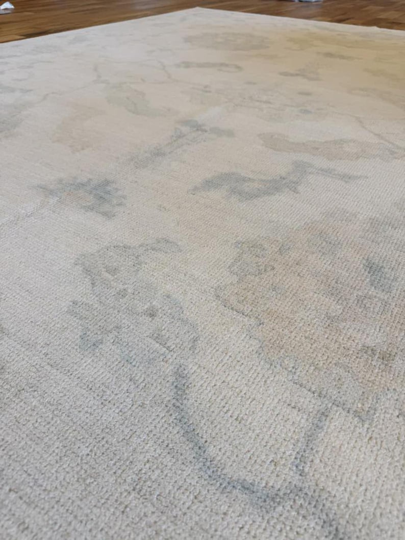 May include: A large area rug featuring a cream-colored base with a subtle, faded pattern in soft shades of light blue and beige. The rug has a low-pile, textured surface with an intricate, repeating design that appears worn or distressed.