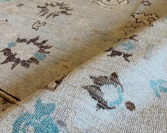 May include: A beige area rug featuring a repeating pattern of stylized floral and geometric motifs in shades of dark brown and light blue. The rug has a textured, woven pile surface with a corner folded over to reveal the underside construction.