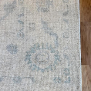 May include: A rectangular area rug featuring a cream-colored base with a faded, light blue floral and geometric pattern. The rug has a textured, woven surface with a visible border design and a soft, low-pile appearance throughout the fabric.
