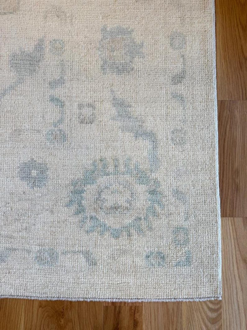May include: A rectangular area rug featuring a cream-colored base with a faded, light blue floral and geometric pattern. The rug has a textured, woven surface with a visible border design and a soft, low-pile appearance throughout the fabric.