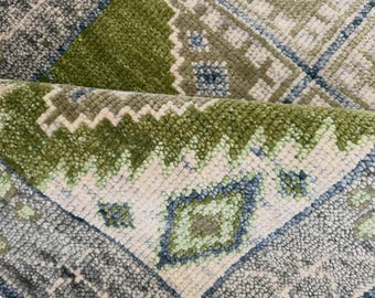 Peut inclure: Un tapis en laine noué à la main présentant un motif géométrique dans des tons de vert olive, sauge, crème et bleu ardoise. Le tapis possède un poil épais et texturé avec un coin replié révélant une frange blanche et un motif en losange.