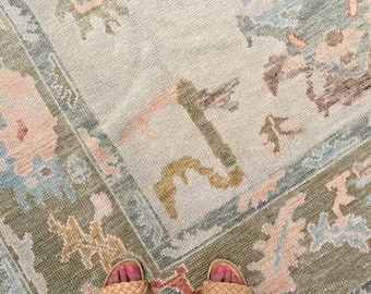 May include: A woven area rug featuring a light cream base with a multi-colored pattern in shades of sage green, dusty pink, light blue, and muted gold. The design includes geometric and floral-inspired motifs arranged in a repeating layout.