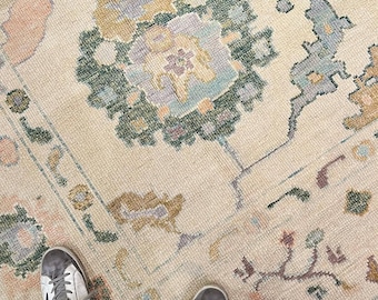 May include: A woven area rug featuring a cream-colored base with a repeating floral and geometric pattern in muted shades of sage green, dusty blue, soft yellow, and pale pink. The design includes stylized blossoms and intricate border motifs.