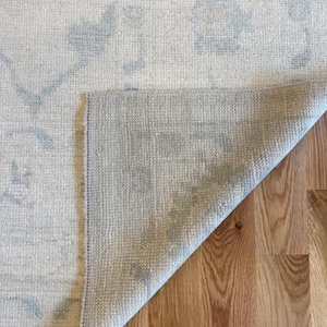 May include: A woven area rug with a cream-colored base and a faded light blue floral pattern. The corner of the rug is folded over to reveal the textured, light-colored underside, showing the dense weave construction of the textile material.