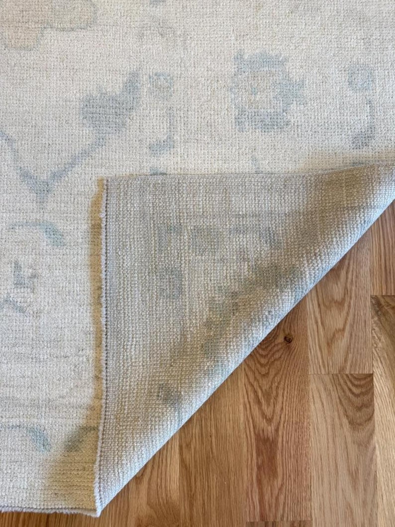 May include: A woven area rug with a cream-colored base and a faded light blue floral pattern. The corner of the rug is folded over to reveal the textured, light-colored underside, showing the dense weave construction of the textile material.