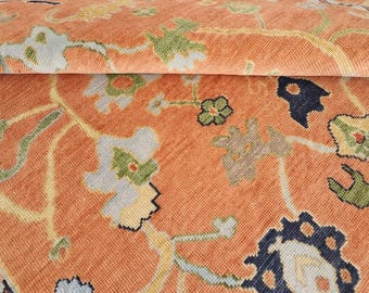 May include: A woven rug featuring a vibrant orange background with a repeating floral pattern in shades of light blue, sage green, cream, and dark navy blue. The textile displays a dense, intricate design with stylized blossoms and vines.