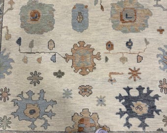 Peut inclure: Un grand tapis avec un fond crème orné d'un motif répétitif de fleurs et de formes géométriques stylisées dans des tons bleu ardoise, orange sourd, vert sauge et gris anthracite, le tout entouré d'une bordure décorative.