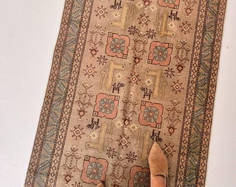 May include: A long, rectangular runner rug featuring a tan background with repeating geometric patterns in muted pink, sage green, and dark brown. The design includes stylized animal motifs and floral shapes, framed by a decorative border.