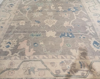 May include: A rectangular area rug featuring a gray field with a central floral motif in shades of blue, cream, and soft pink. The rug has a wide cream-colored border with a repeating geometric and floral pattern, finished with a thin blue edge.
