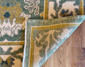 Peut inclure: Un tapis tissé présentant un motif floral et géométrique détaillé dans des tons de vert sauge, jaune moutarde, crème et bleu ardoise. Le tapis est plié à un coin pour révéler le dessous, montrant le poil dense et la structure tissée.