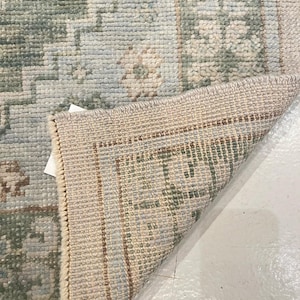 May include: A woven area rug featuring a light blue and sage green floral pattern with a cream-colored border. The corner of the rug is folded over to reveal the textured, light-colored underside and the dense, knotted construction of the pile.