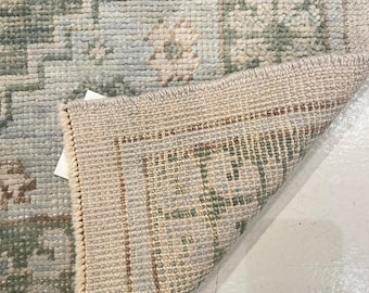 May include: A woven area rug featuring a light blue and sage green floral pattern with a cream-colored border. The corner of the rug is folded over to reveal the textured, light-colored underside and the dense, knotted construction of the pile.