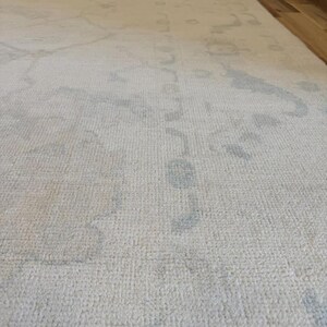 May include: A large area rug featuring a cream-colored base with a subtle, faded pattern in light gray. The rug has a low-pile, woven texture and covers a significant portion of the floor, displaying intricate, soft-edged floral and geometric motifs.