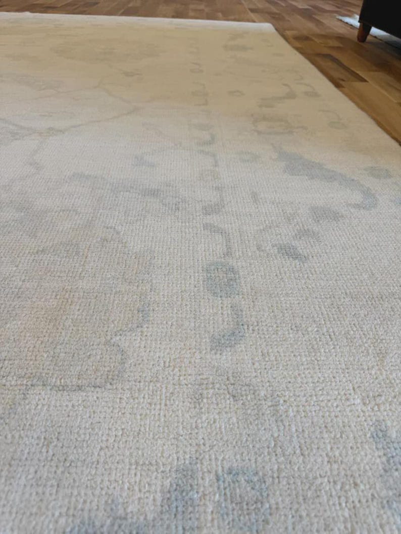 May include: A large area rug featuring a cream-colored base with a subtle, faded pattern in light gray. The rug has a low-pile, woven texture and covers a significant portion of the floor, displaying intricate, soft-edged floral and geometric motifs.