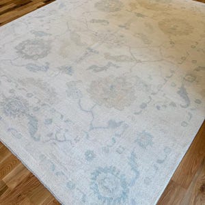 May include: A large rectangular area rug featuring a cream-colored base with a faded, intricate floral pattern in soft shades of light blue and beige. The design includes symmetrical botanical motifs and scrolling vines across the surface.