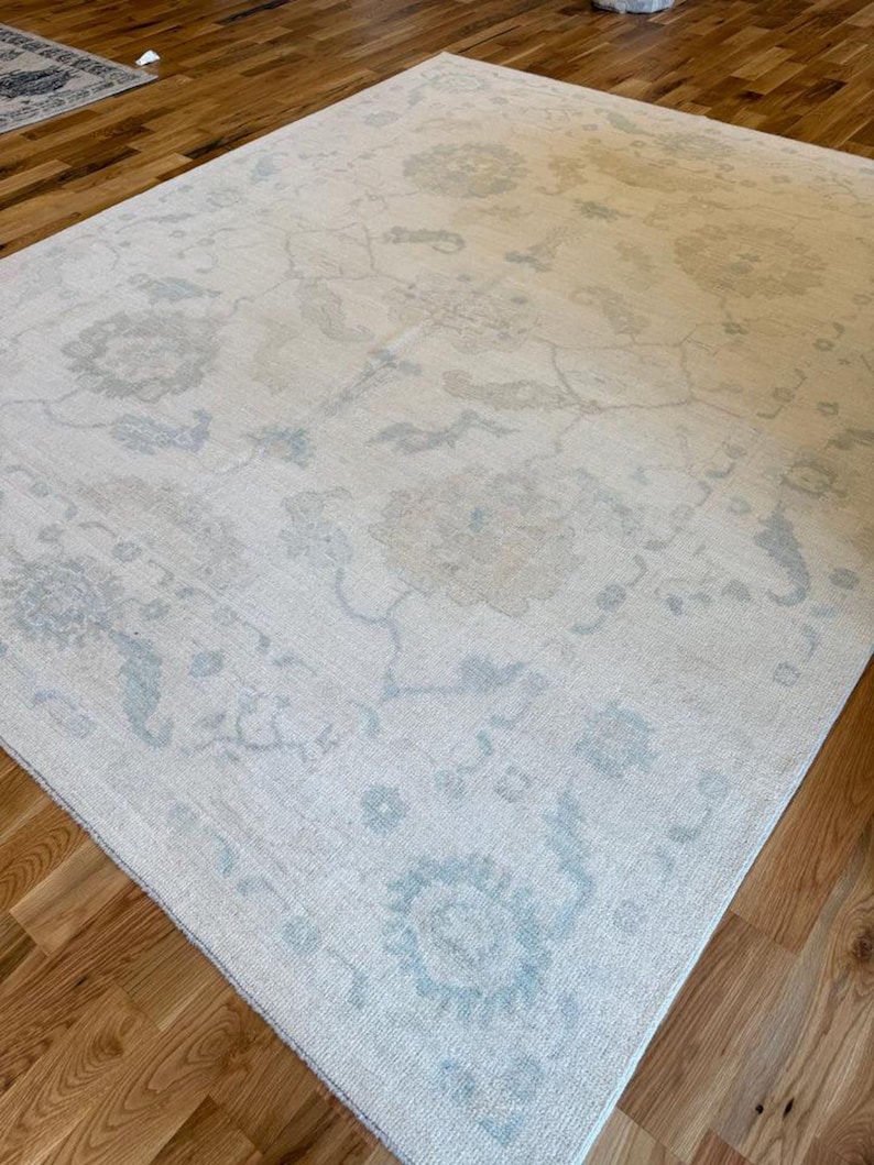 May include: A large rectangular area rug featuring a cream-colored base with a faded, intricate floral pattern in soft shades of light blue and beige. The design includes symmetrical botanical motifs and scrolling vines across the surface.